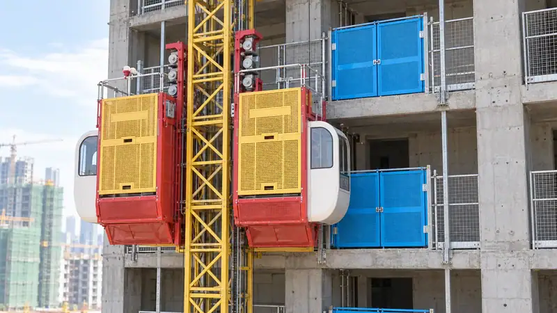 Construction lift with blue safety barriers on a multi-story building under construction, featuring a yellow crane in the background.