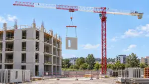 Tower crane lifting a concrete panel at a construction site with a partially built multi-story building and clear blue sky.