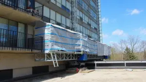 Plateforme de travail grimpante à mât unique IHURMO enveloppée d’une bâche de protection contre les intempéries, le long de la façade d’un immeuble résidentiel